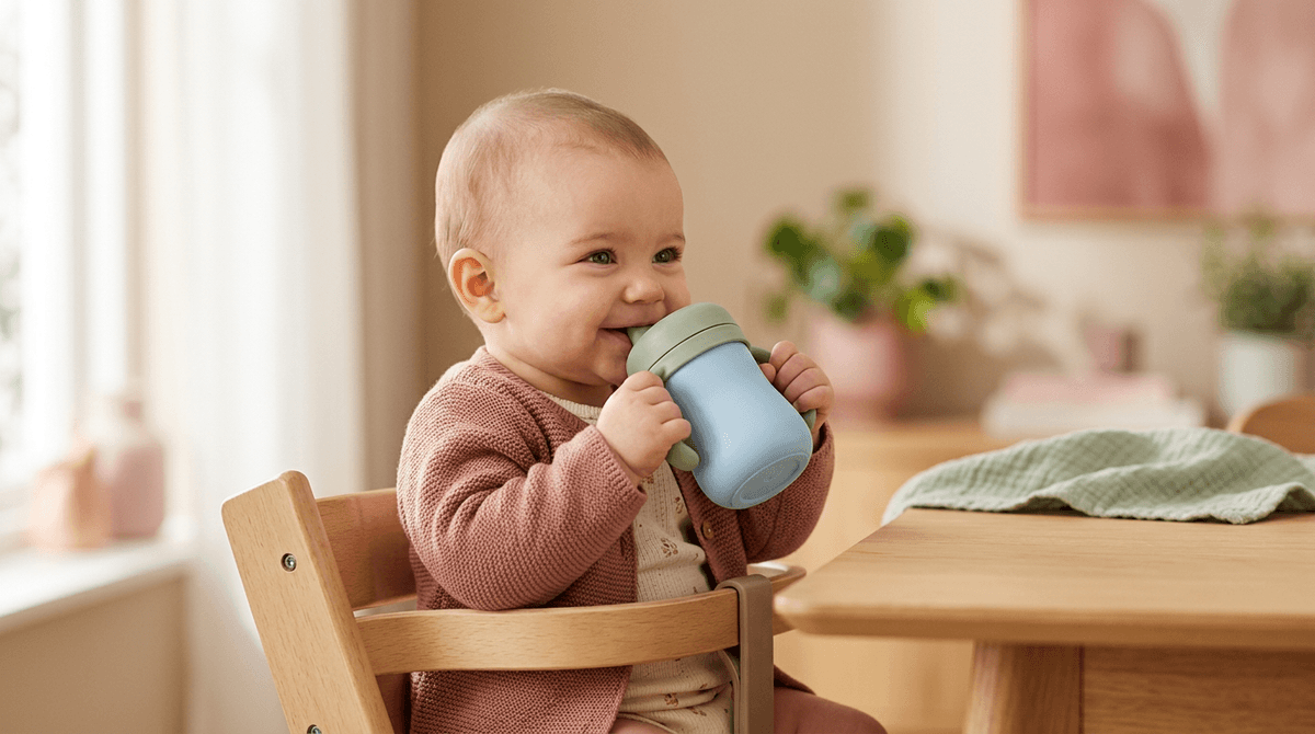 Bébé souriant buvant de l'eau dans une tasse à anses, assis dans sa chaise haute.